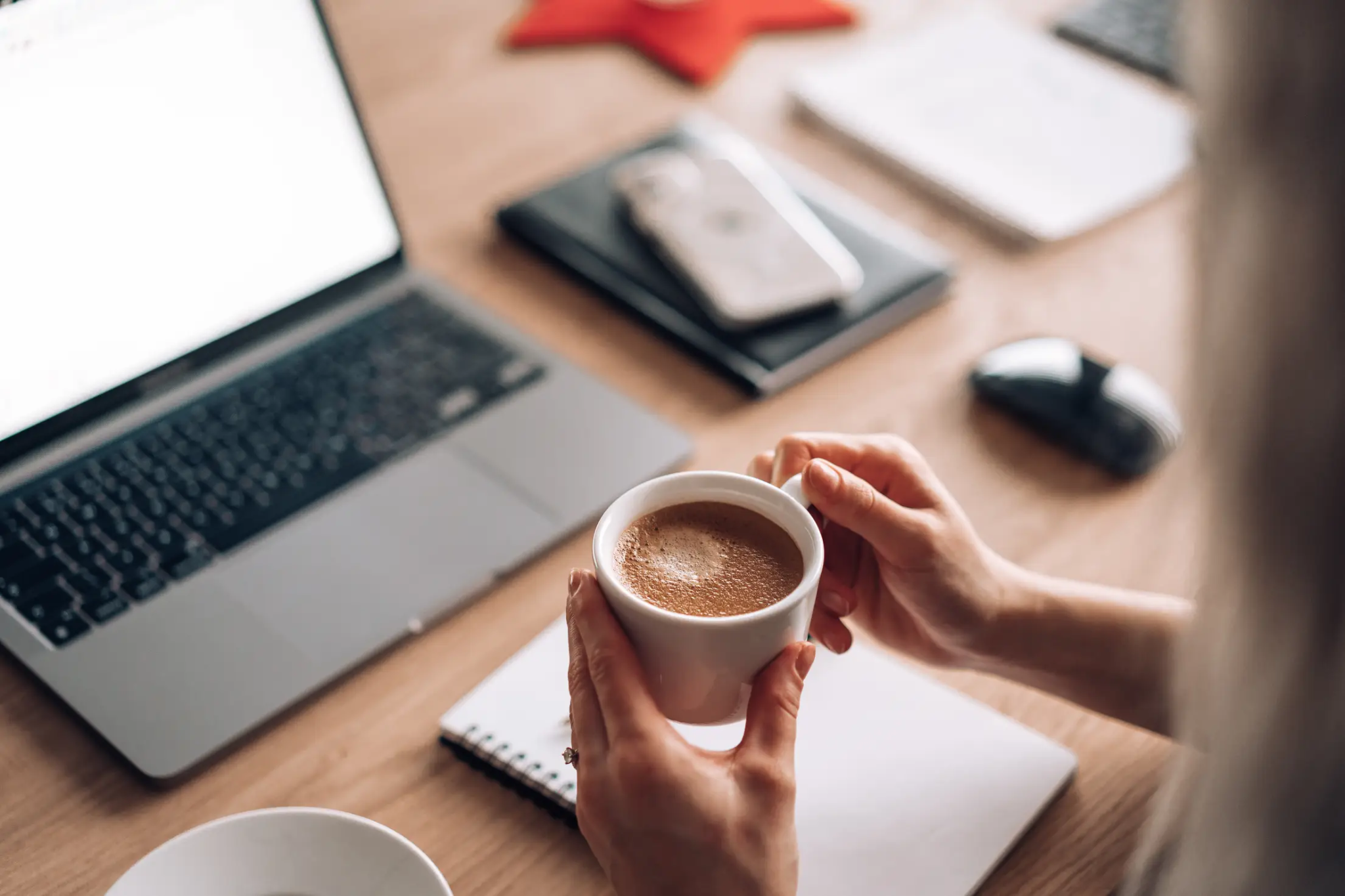 Person holding a cup of coffee at a desk with a laptop, notebook, phone, and household paperwork